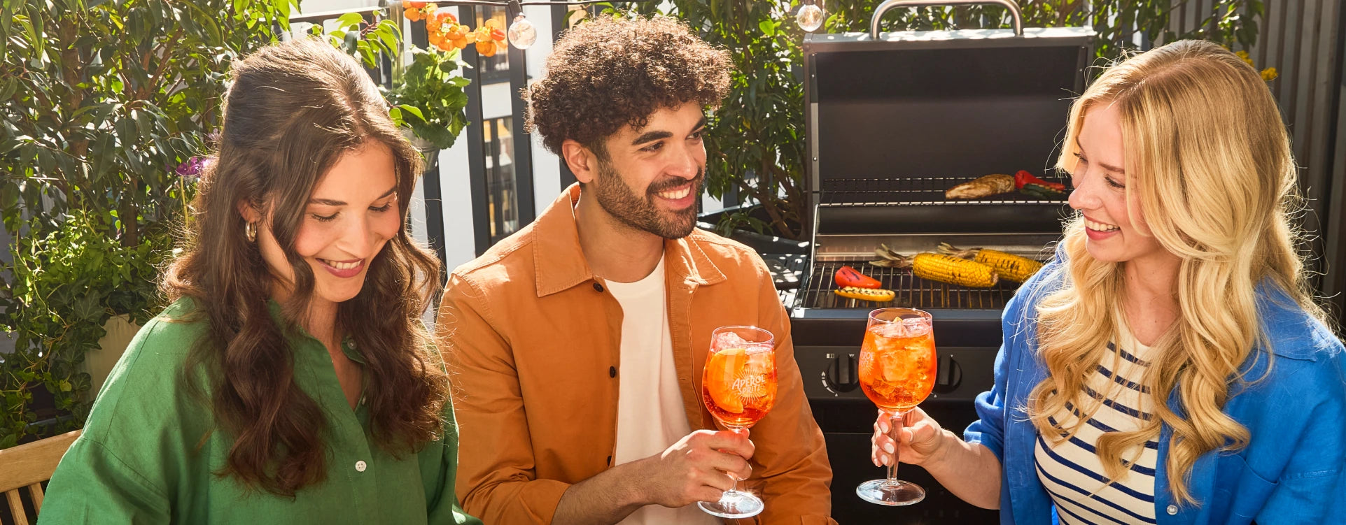 Three friends enjoying Aperol Spritz cocktails while sitting by a barbecue grill with grilled vegetables